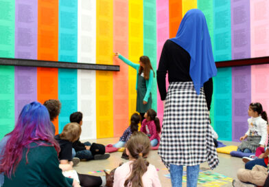 Jugendliche Personen in einer Ausstellung vor einer Wand mit bunten Plakaten, eine Person im Hintergrund erklärt das Kunstwerk
