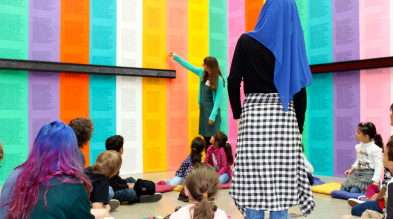 Jugendliche Personen in einer Ausstellung vor einer Wand mit bunten Plakaten, eine Person im Hintergrund erklärt das Kunstwerk