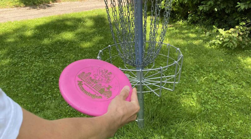 Der Arm einer Person mit einer Frisbee-Scheibe, die auf einen Korb aus Metallketten zielt