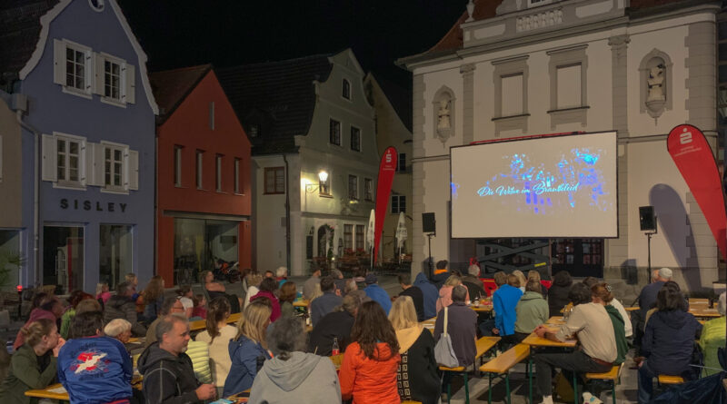 Auf dem Theaterplatz sitzen im Dunkeln Menschen an Bierbänken und schauen Kurzfilme die auf eine Leinwand am Theatergebäude projiziert werden