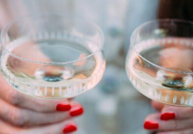 Nahaufnahme von zwei Händen mit rot lackierten Fingernägeln, die jeweils ein gefülltes Champagnerglas halten