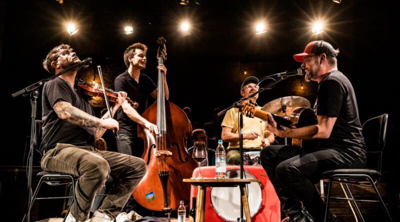 Die Band Django3000 sitzt mit Instrumenten im Kreis auf einer Bühne